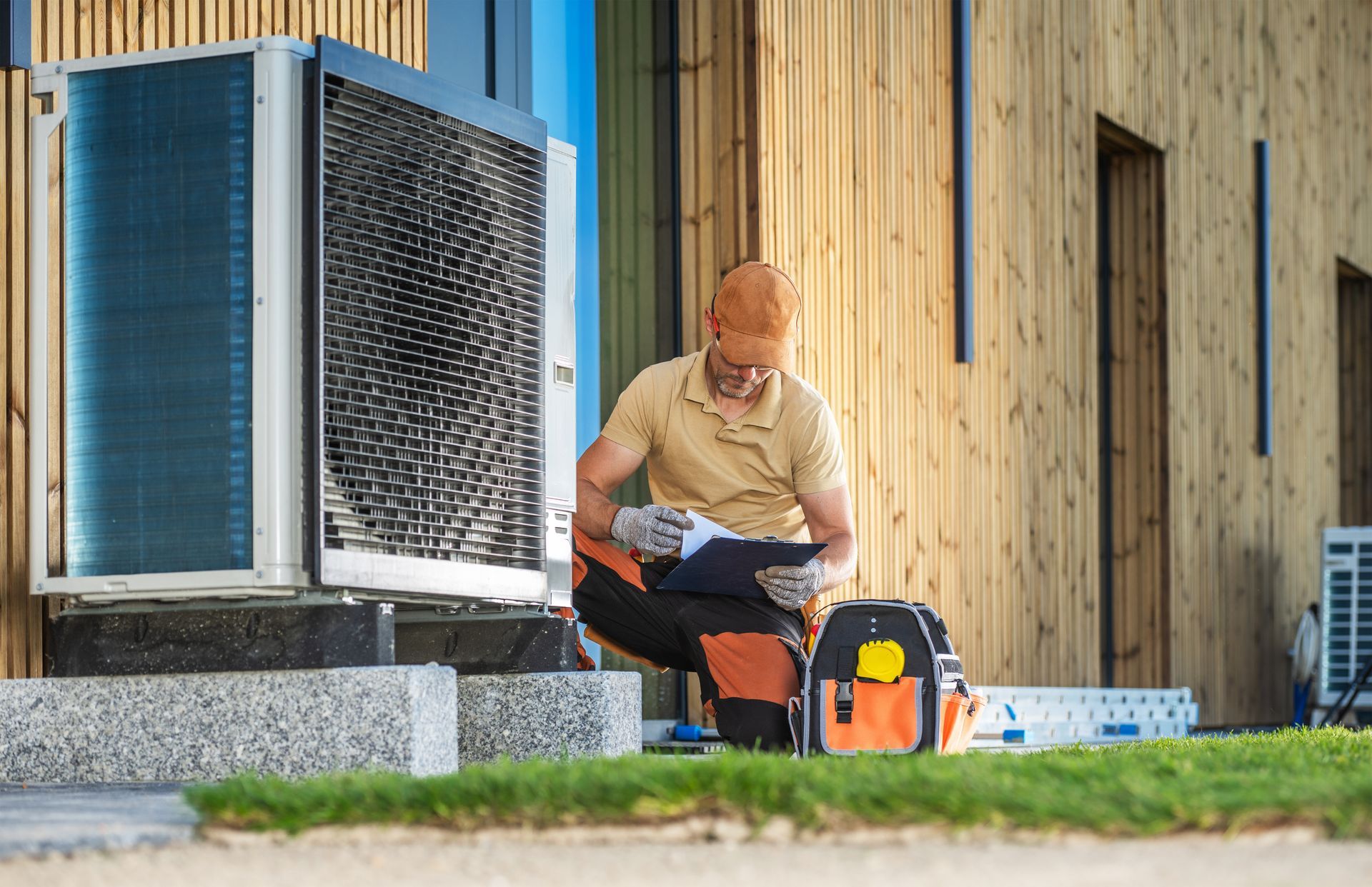 Man kneels near an HVAC unit, inspecting paperwork. Orange tool bag beside him, wooden building in background.