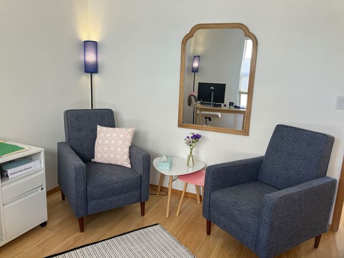 Two blue armchairs face each other with a small table, rug, and mirror in a brightly lit, minimalist office space.