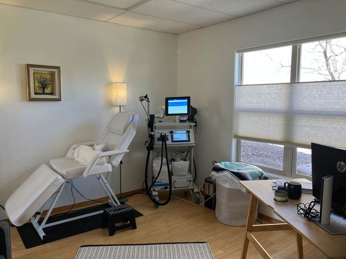 A small, bright treatment room with a white facial chair, a laser equipment cart, and a desk in front of a window.