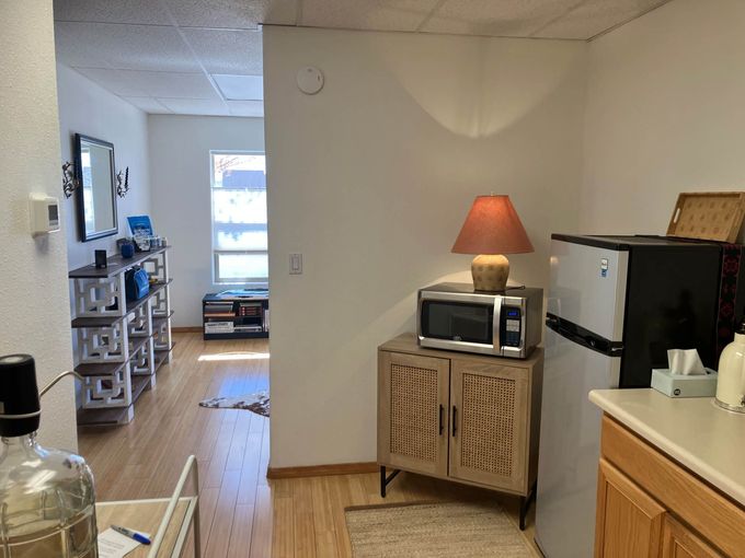 An interior view of a room with light wood floors, a wooden cabinet with a microwave and lamp, and a refrigerator.