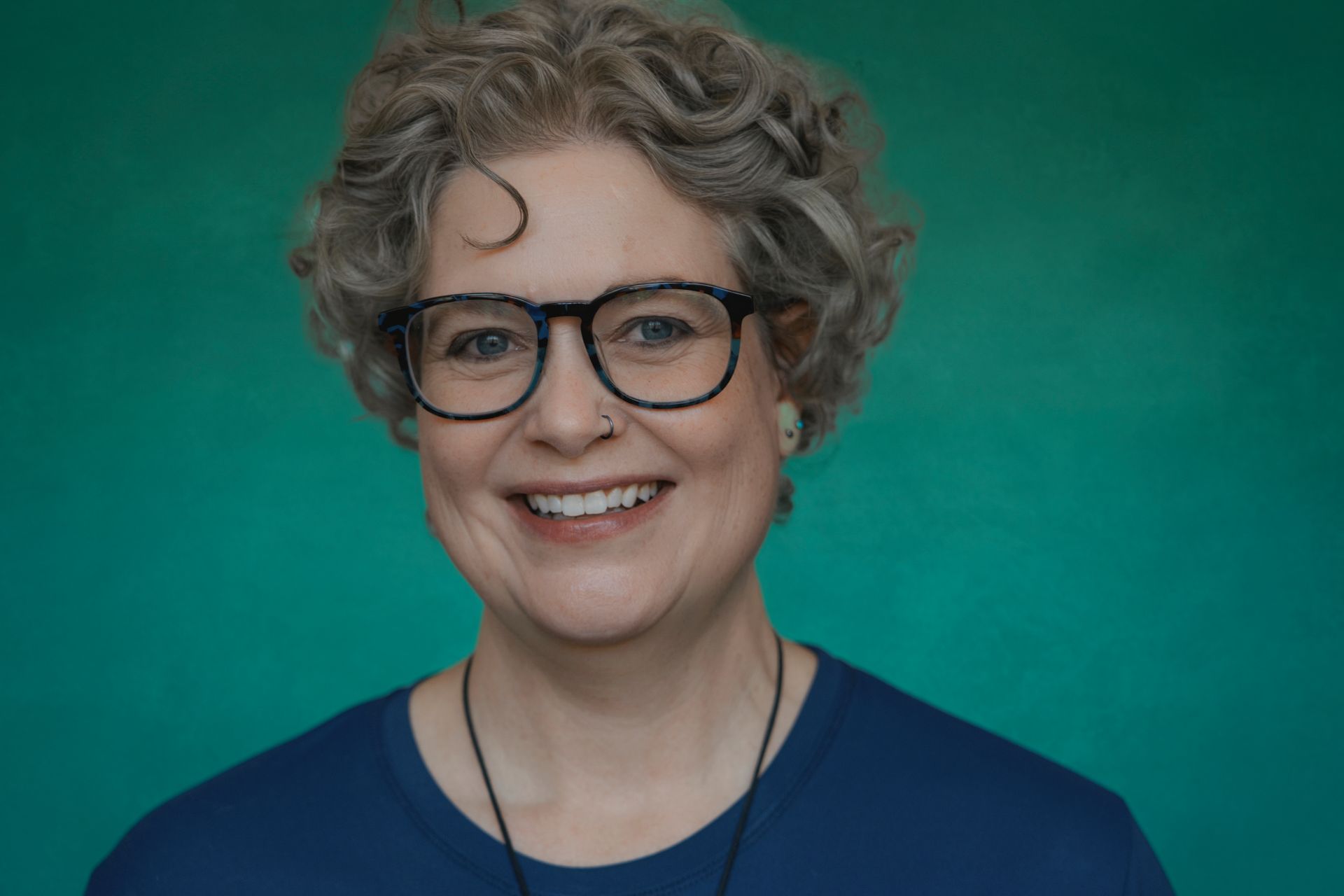 Rachel Joyce with curly gray hair, glasses, and a blue shirt smiles against a teal background.