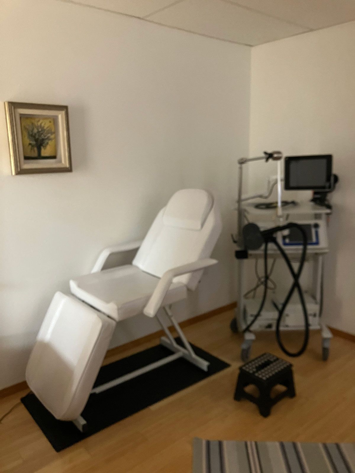 White medical exam chair in a corner of a room, with a machine, a stool, and a small painting.
