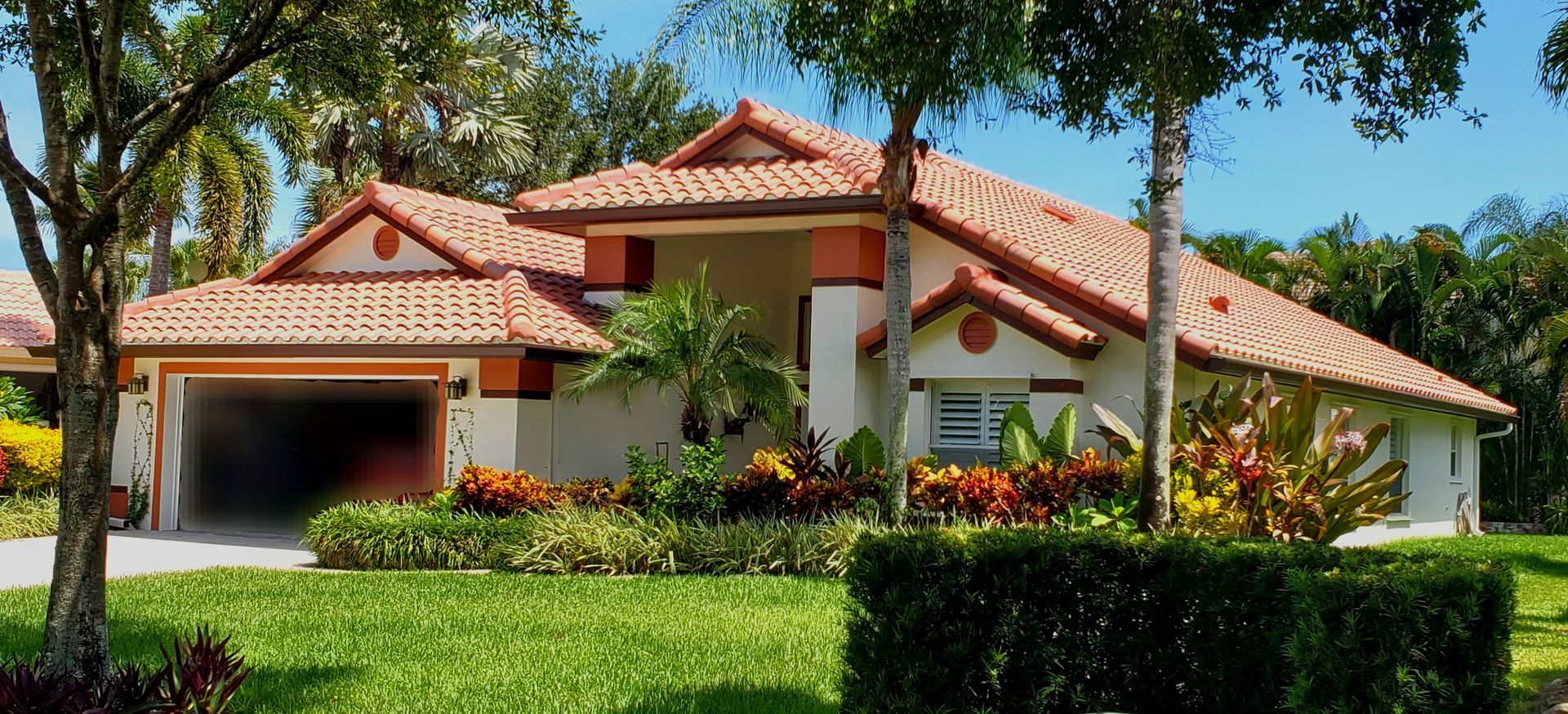 A large white house with a red tile roof is surrounded by trees and bushes.