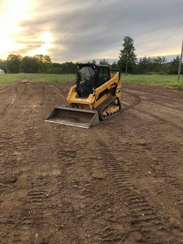 A yellow tracked skid steer loader sits on a large, cleared dirt field under a cloudy sky at sunset.