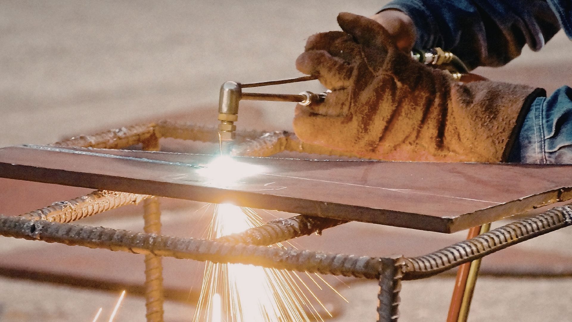 A person wearing heavy leather gloves uses a torch to cut through a thick metal plate resting on a rebar frame.