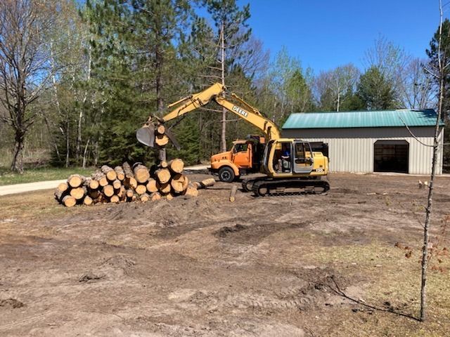 Yellow excavator lifting a log onto a stack beside a truck and metal building on a sunny day.