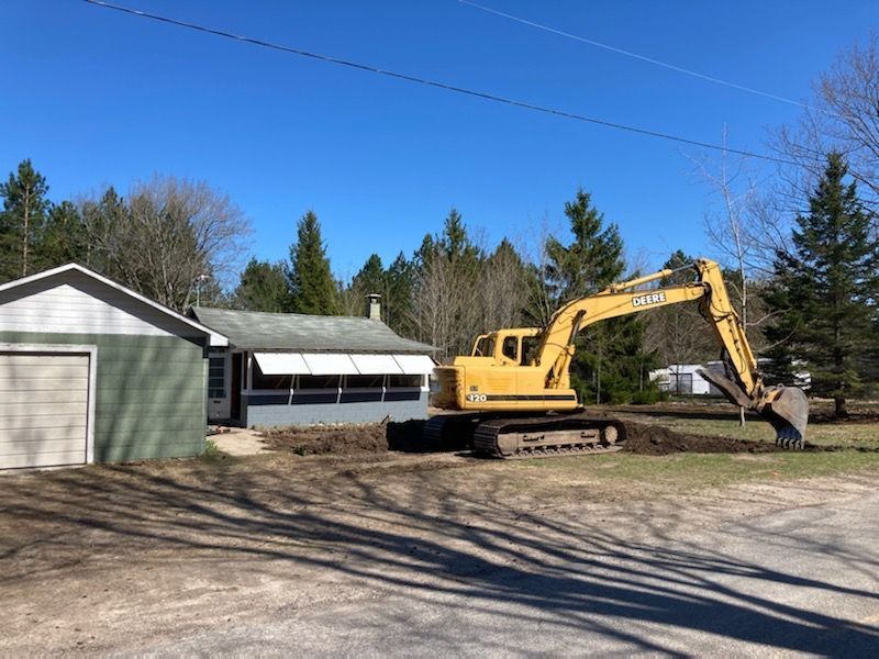 A yellow excavator sits in the yard of a small house with a detached garage on a sunny day.