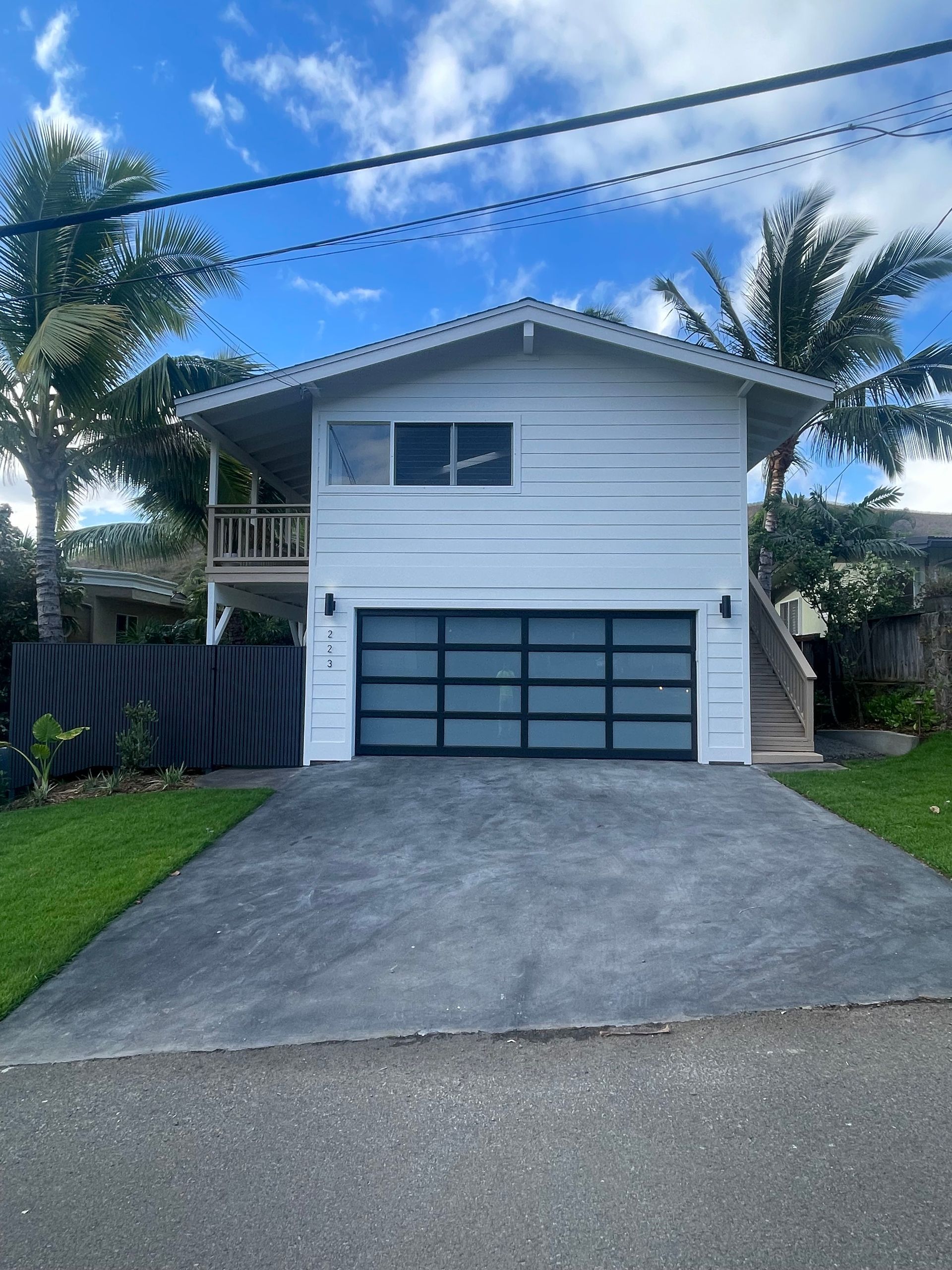 White two-story house with a gray driveway, glass garage door, and palm trees against a blue sky.