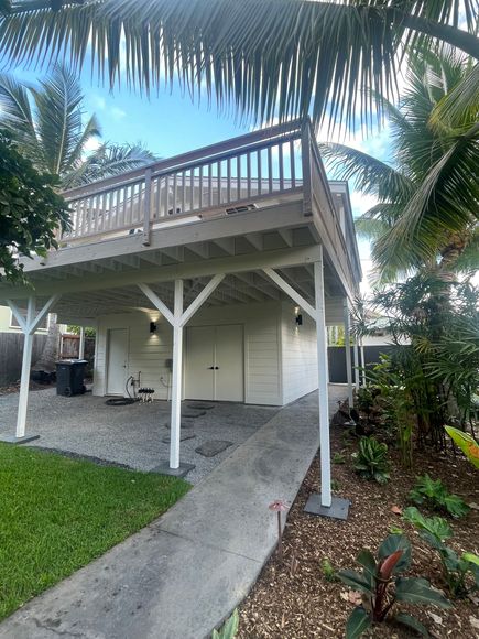 Elevated white building with deck; driveway in front. Lush green grass and plants surround. Blue sky.