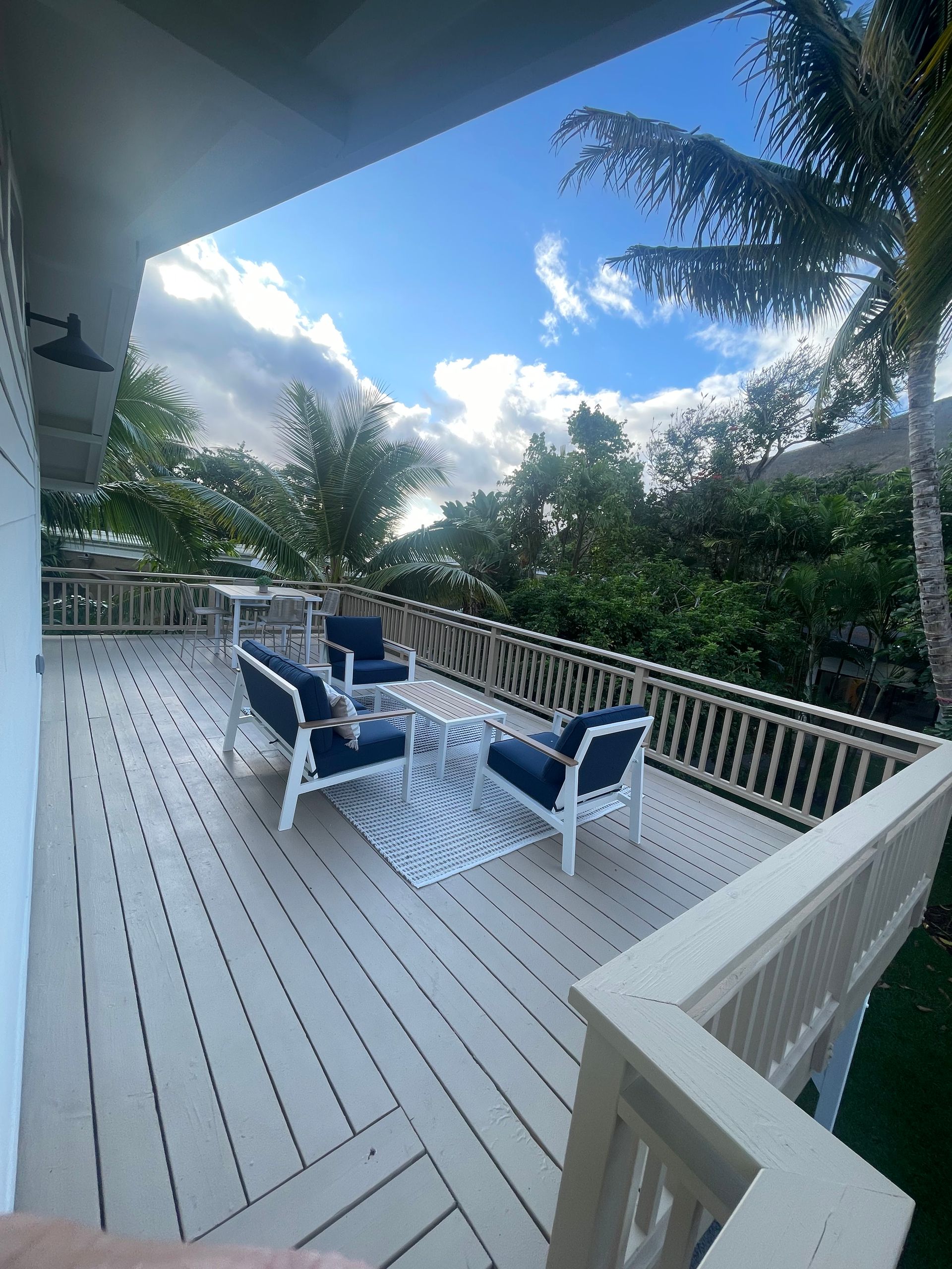 Deck with outdoor seating, blue chairs, white rug, and lush green trees under a bright blue sky.