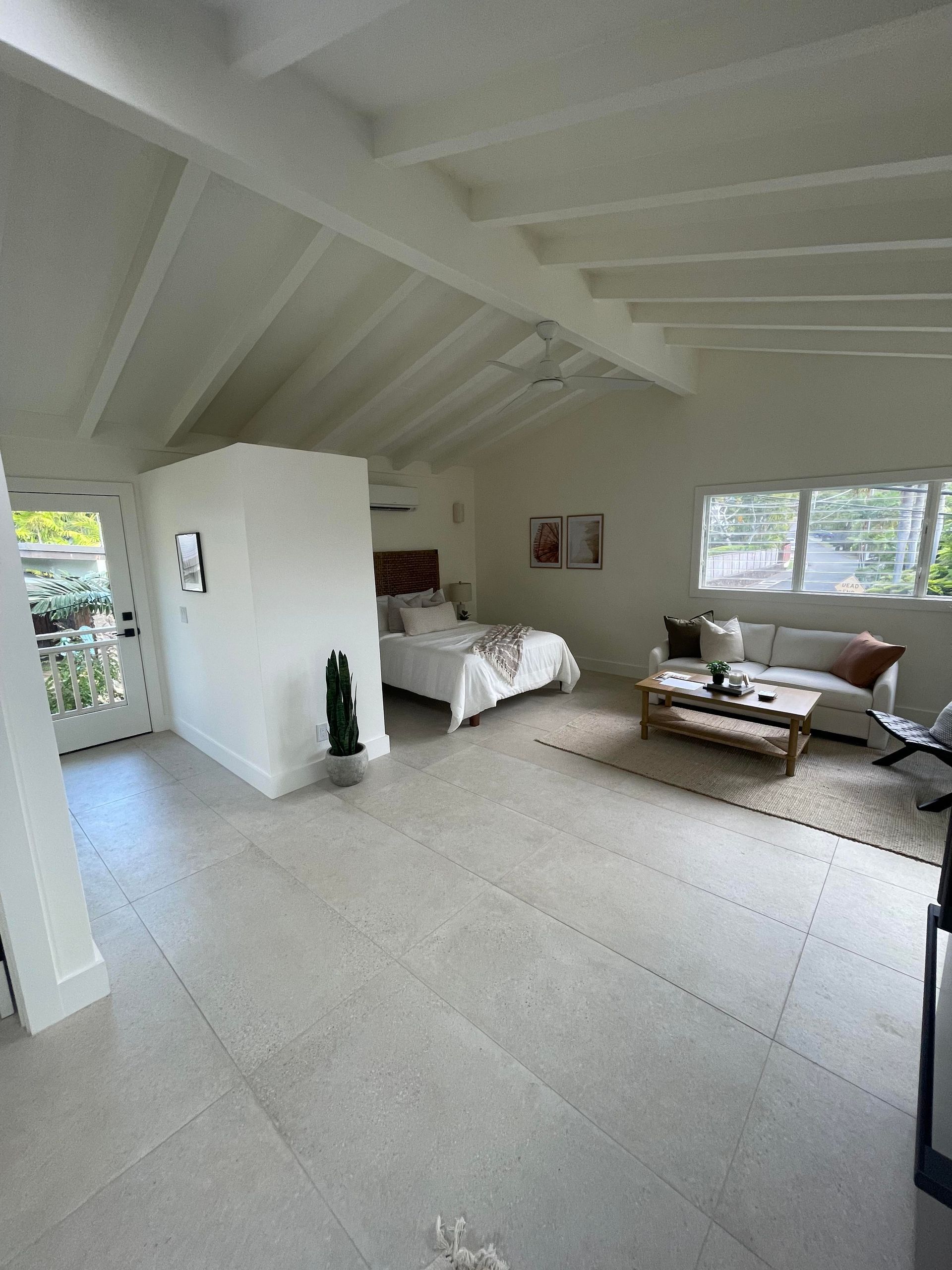 Spacious, bright room with a bed, sofa, and neutral-colored tile floor. White walls and exposed beams.