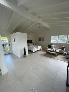Spacious, bright room with a bed, sofa, and neutral-colored tile floor. White walls and exposed beams.