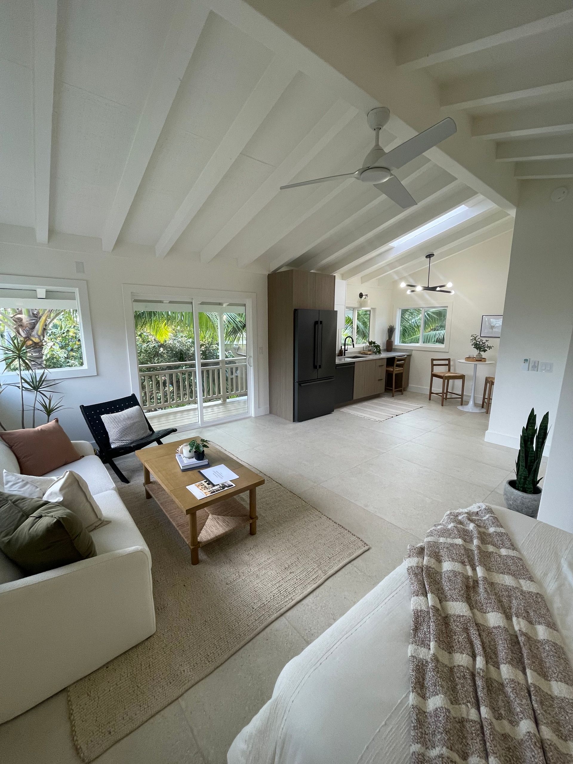 Living room with vaulted ceiling, windows, white sofa, wooden coffee table, and kitchen visible in the background.
