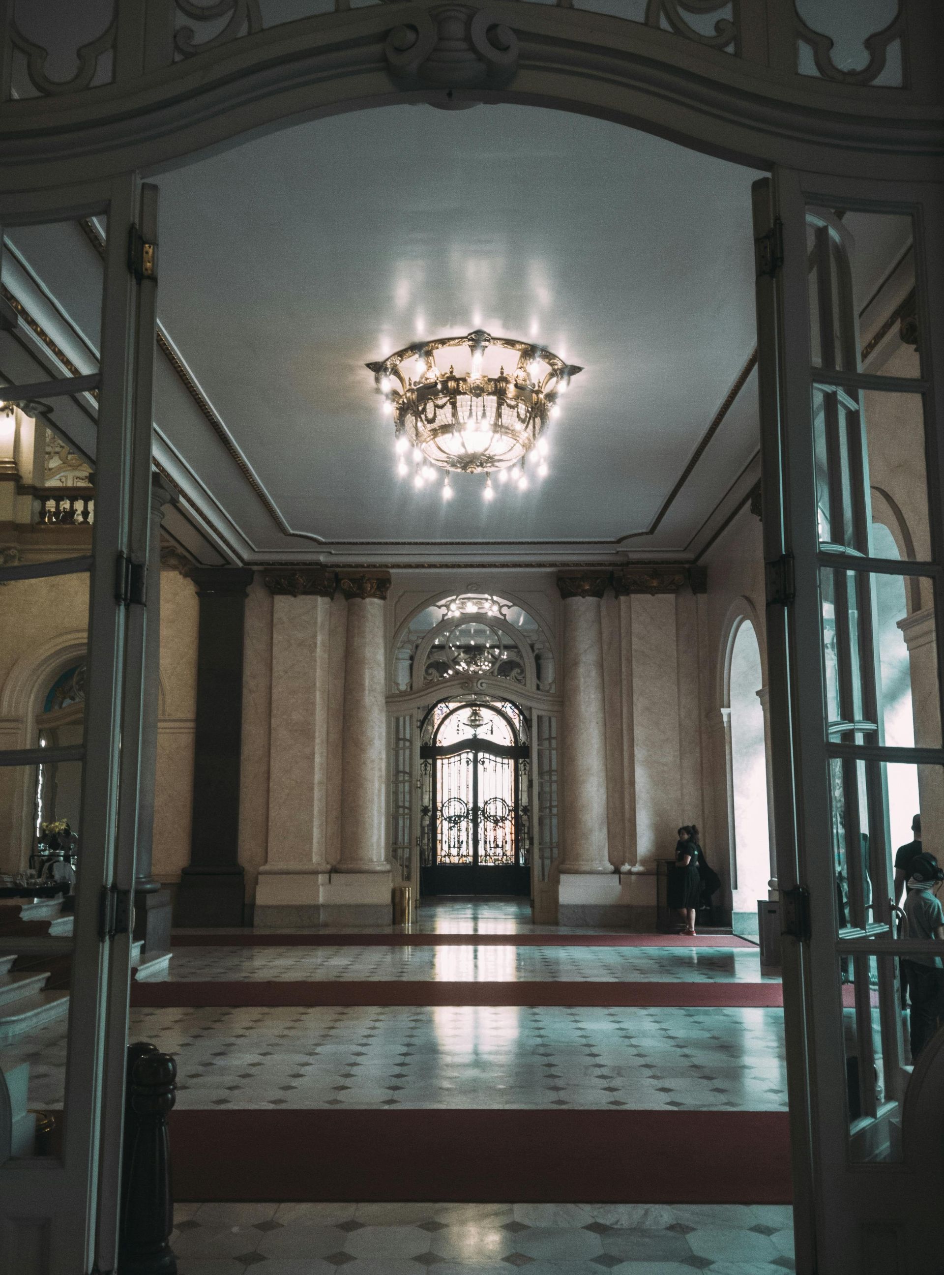 Gran salón con lámpara de araña ornamentada, pisos de mármol y puertas dobles abiertas.