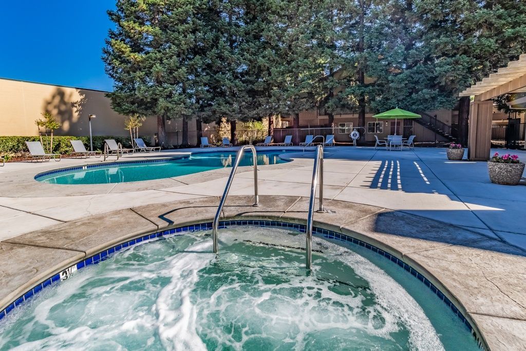 Outdoor pool and hot tub area at an apartment complex with lounge chairs and trees.