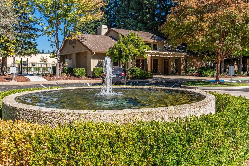 Circular stone fountain in front of a beige apartment building with trees and shrubs.