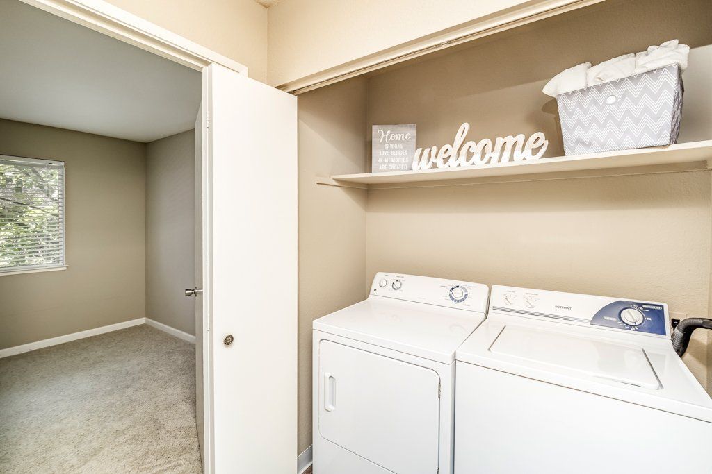 Laundry closet with a washer and dryer side by side, shelf above, basket, and decorative sign.