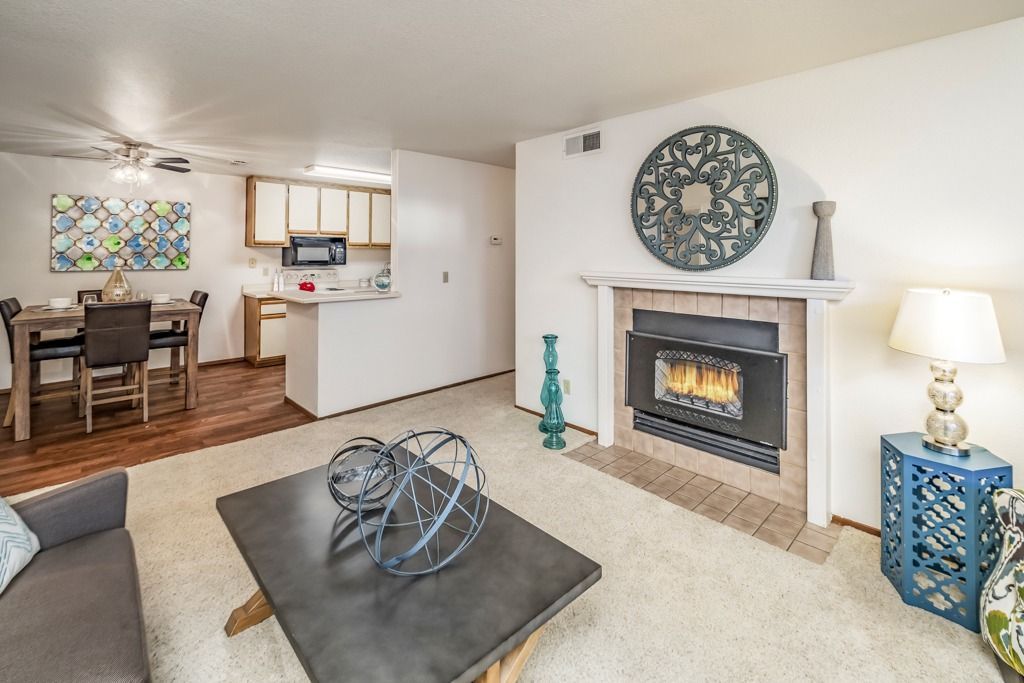 Living room with a gas fireplace, beige carpet, and open kitchen in the background.