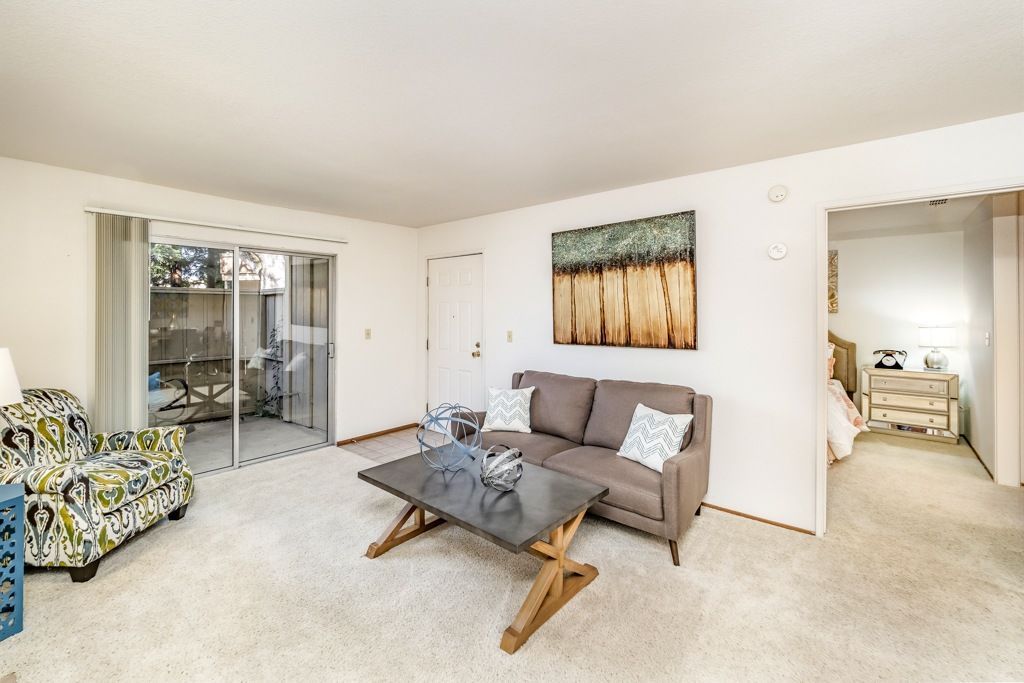 Living room of an apartment with a gray sofa, patterned accent chair, coffee table, and sliding glass door to a patio.