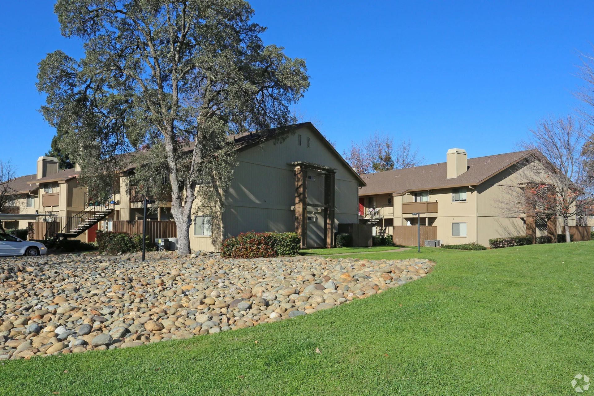 Exterior view of a beige apartment complex with lush lawn, rock landscaping, and a large tree.