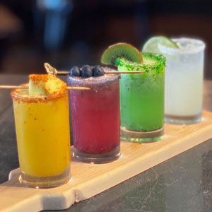 Four colorful cocktails on a wooden tray: yellow, red, green, and white, each garnished with fruit.