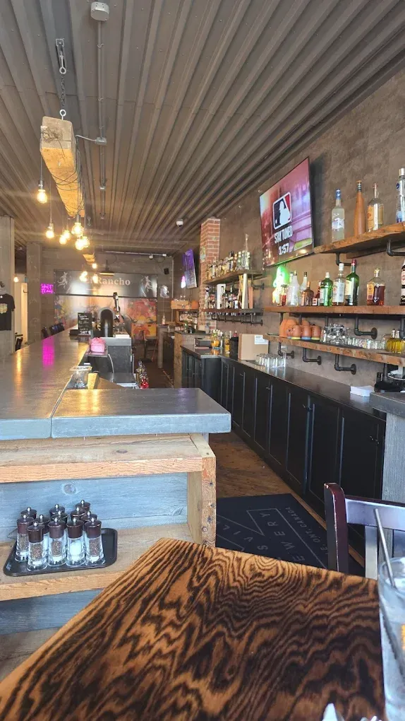 Interior view of a bar with a long concrete counter, wooden accents, and shelves stocked with bottles.