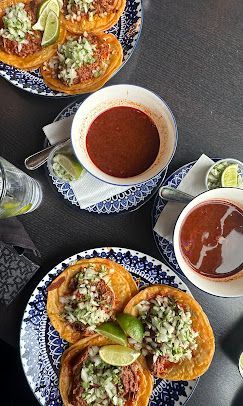 Tacos on blue and white plates, two bowls of red sauce, lime wedges, and a water bottle on a dark table.
