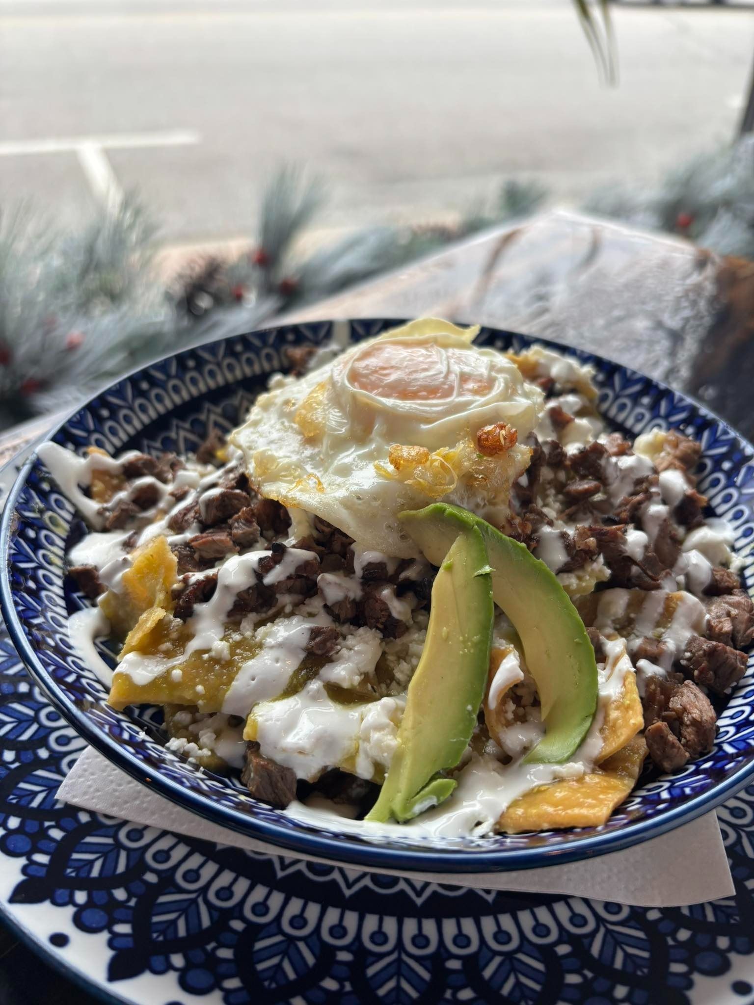 Dish of chilaquiles with egg, avocado, and white sauce in a blue patterned bowl.