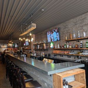 Interior view of a bar with a long concrete counter, shelves of liquor, and a TV screen.