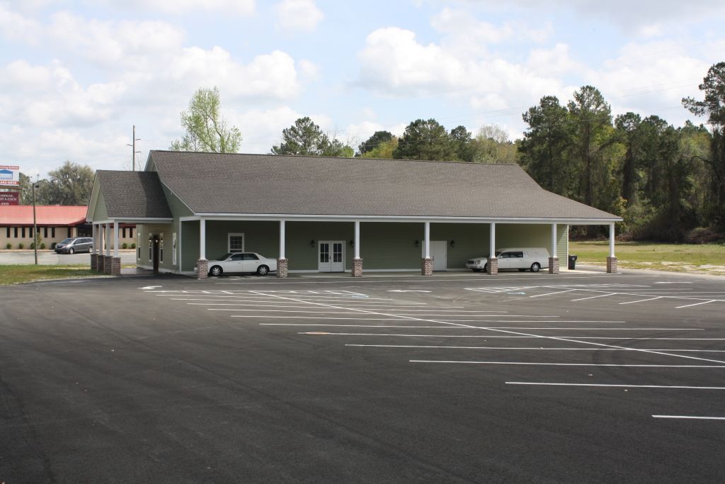 A light green building with a porch and a large asphalt parking lot. A car and a hearse are parked there.