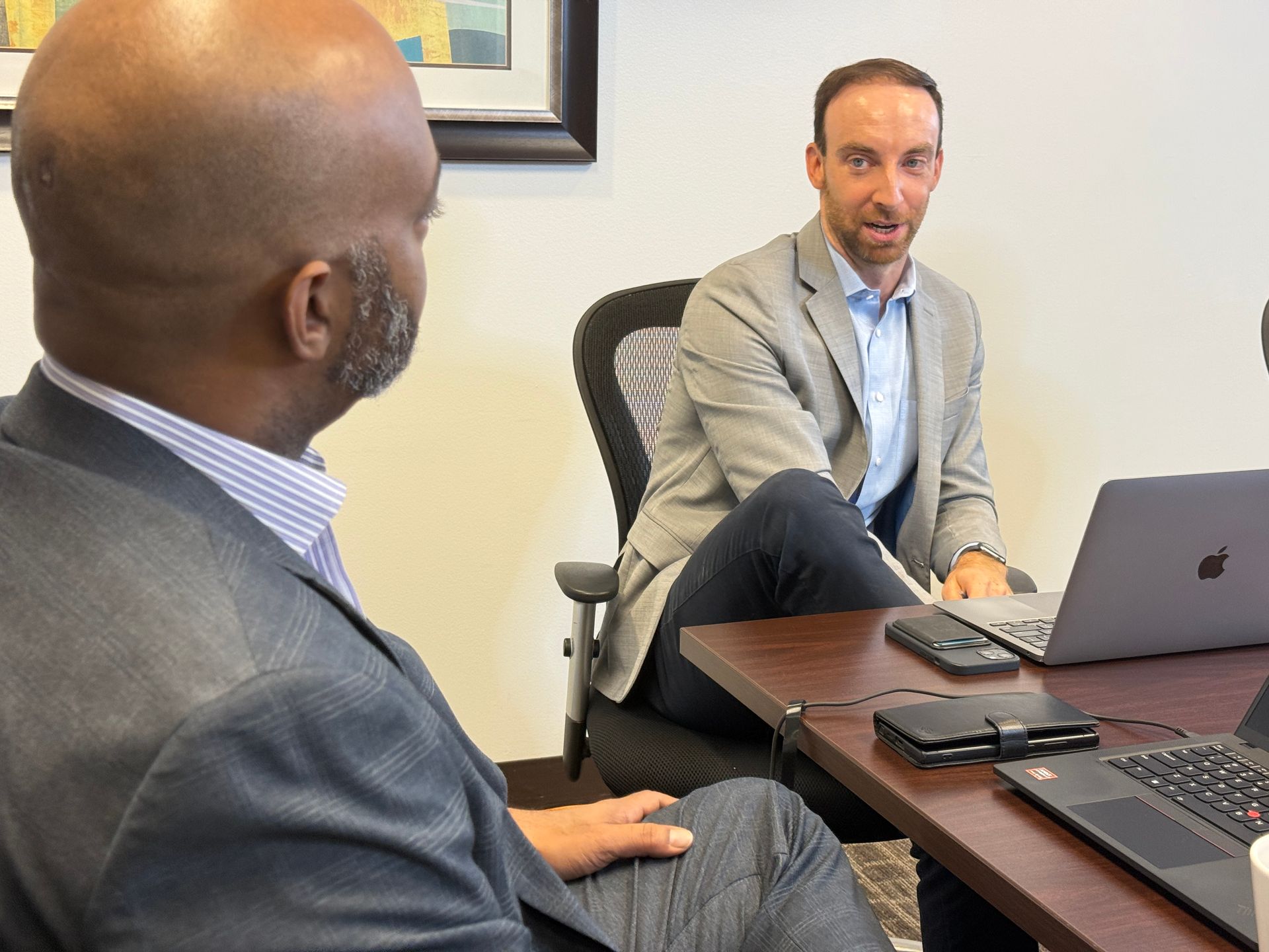 Two men in an office meeting across a desk with laptops open, one speaking while seated in a chair.