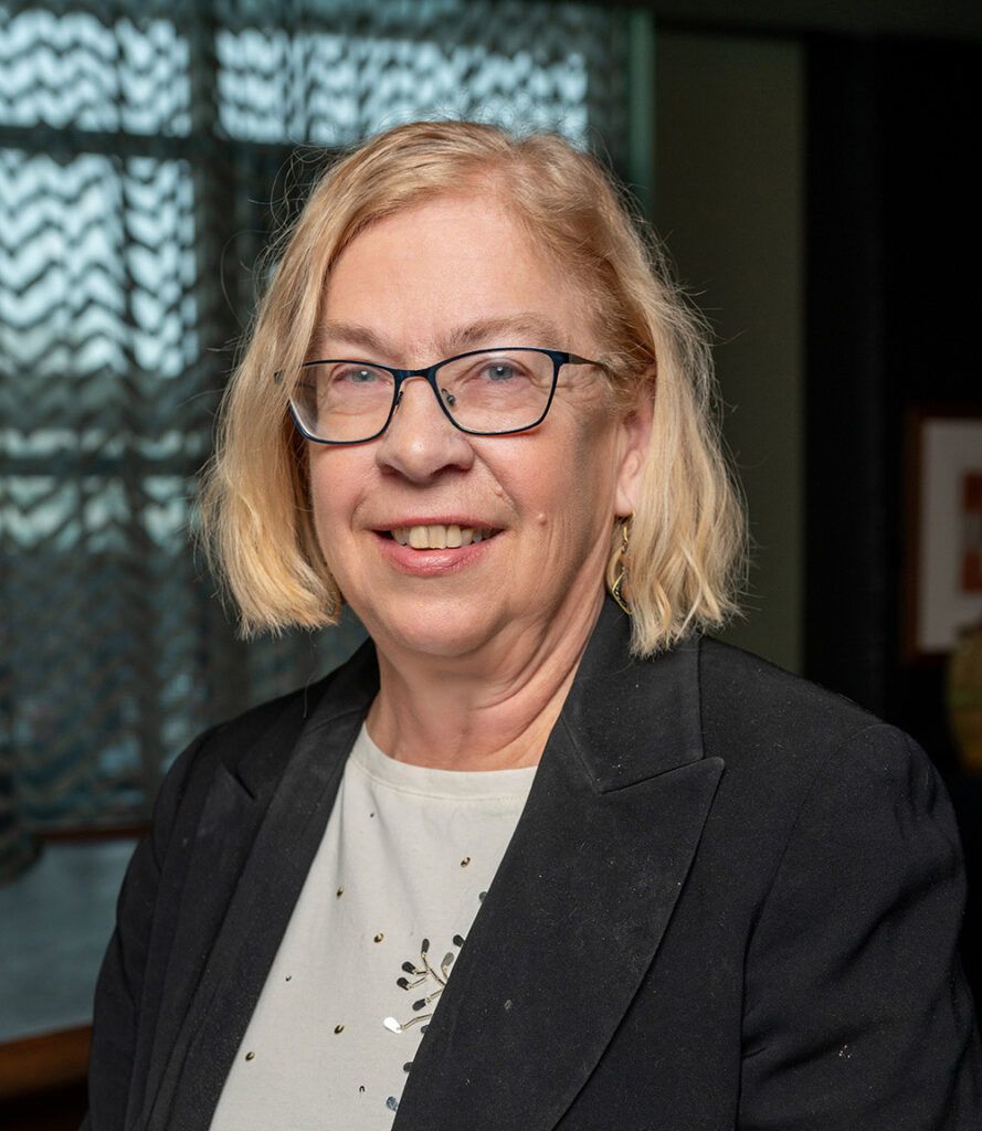 Woman with blonde hair, glasses, and a black blazer smiles in an indoor setting.