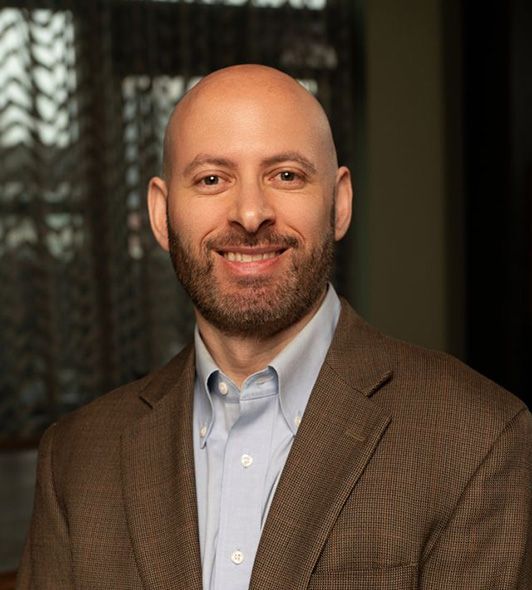 Smiling man with a bald head and beard, wearing a blue shirt and brown blazer.