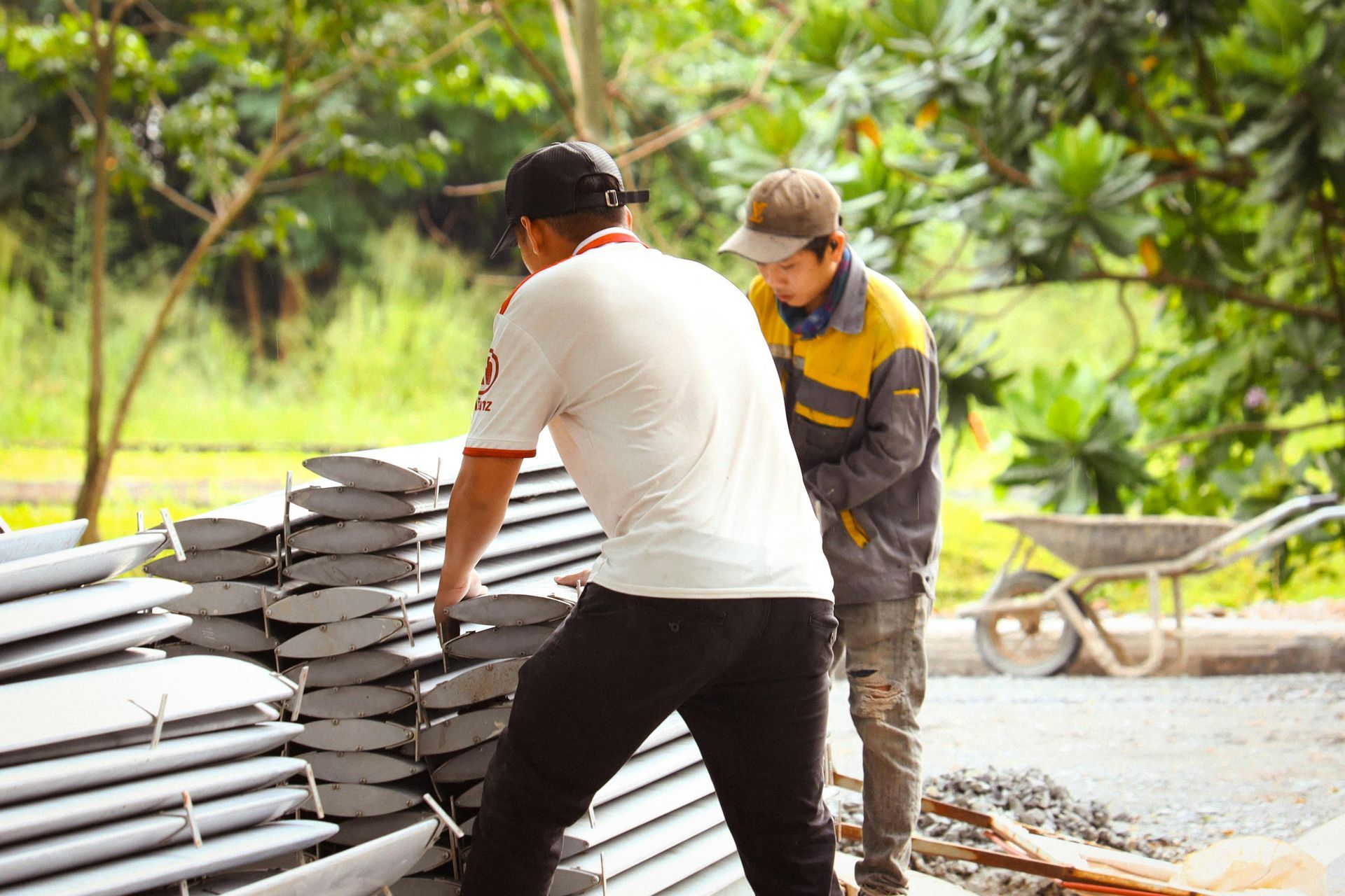 Two people lifting gray metal panels outdoors near construction materials.