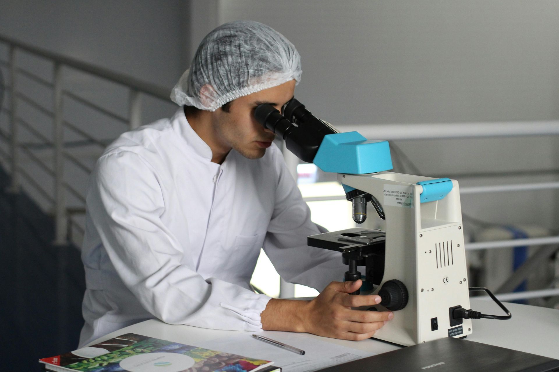 Man in lab coat and hair net looking into a microscope in a lab.