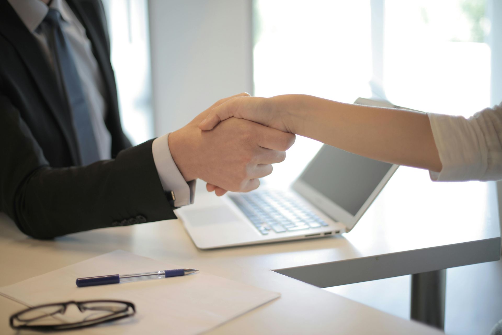 Man in suit shaking hands with a person, seated at a desk with a laptop and pen.