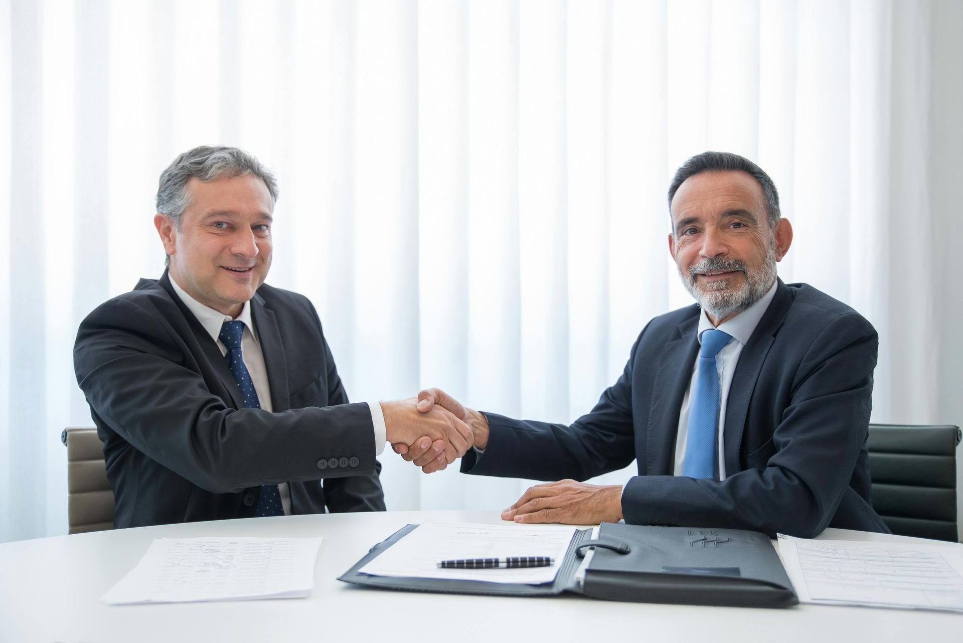 Two businesspeople shaking hands across a desk with documents and a laptop in a bright office