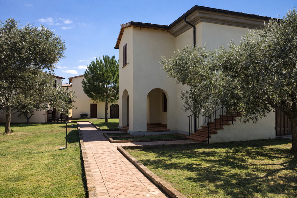 Edifici beige a due piani con ingressi ad arco, vialetti in mattoni, ulivi e prato verde sotto il cielo azzurro.