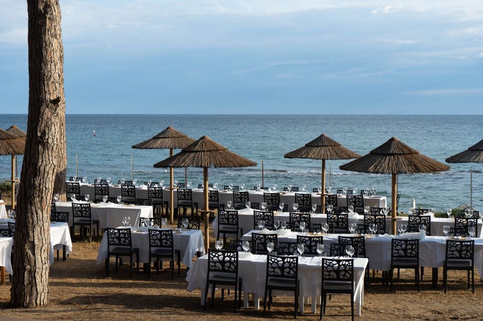 Tavoli apparecchiati per un evento sulla spiaggia, sotto ombrelloni di paglia, di fronte all'oceano.
