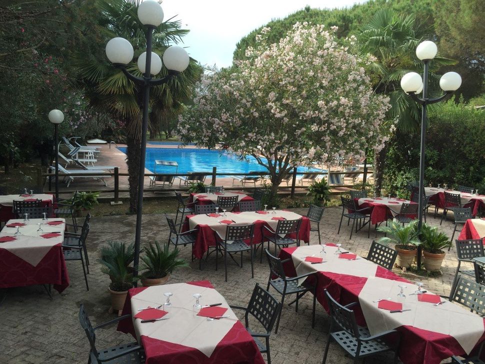 Tavoli da ristorante all'aperto apparecchiati per cenare vicino a una piscina, alberi rigogliosi e lampioni decorativi.