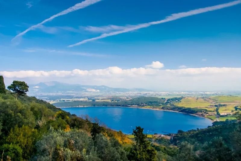 Lago blu con colline verdi, sotto un cielo azzurro con nuvole bianche e scie di condensazione. Città visibile in lontananza.