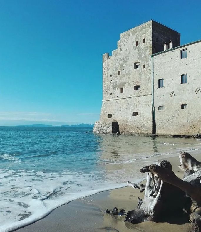 Vista costiera: torre in pietra su una spiaggia, onde che si infrangono sulla riva sotto un cielo azzurro.