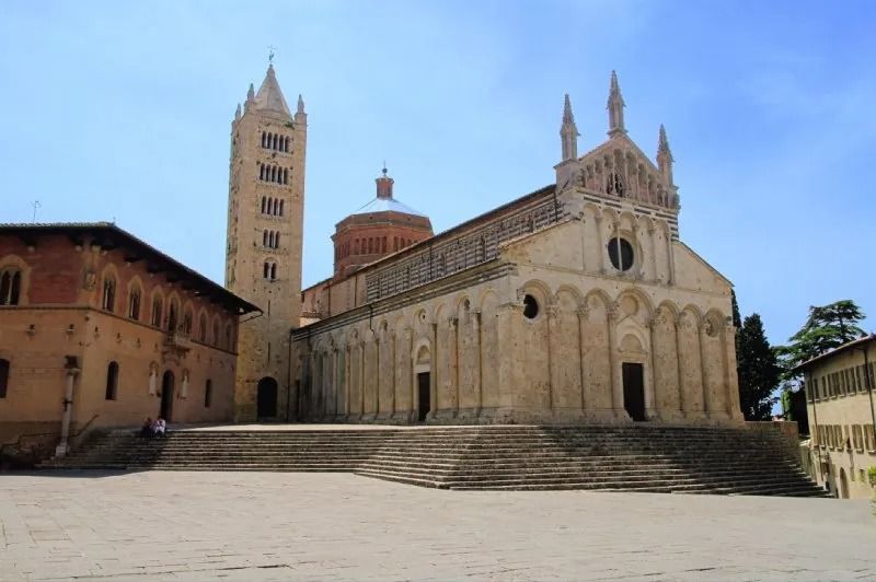 Una grande cattedrale in pietra con campanile e gradini in una piazza sotto un cielo azzurro.