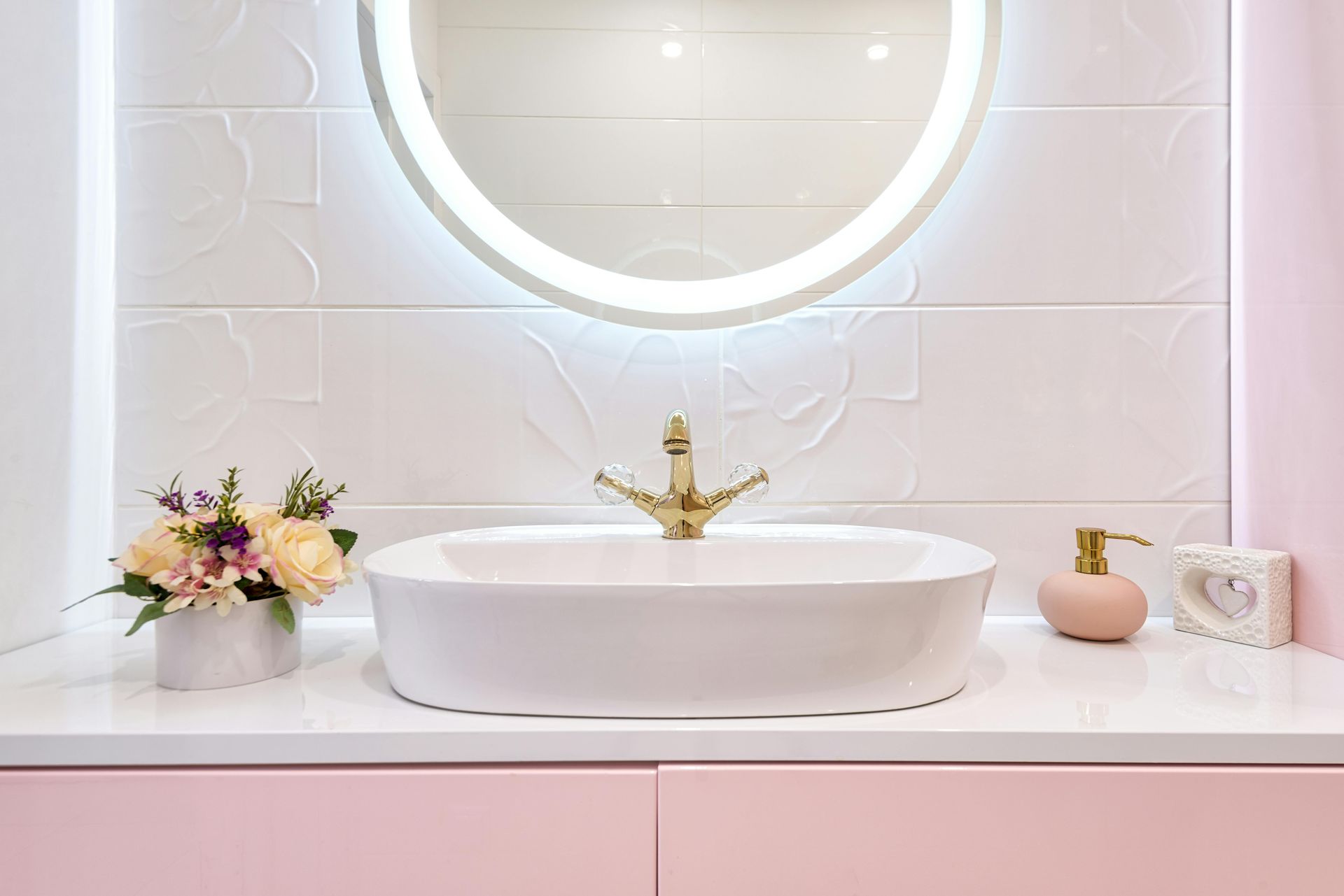 A modern bathroom vanity with a white oval sink, gold faucet, round illuminated mirror, and pink accents.