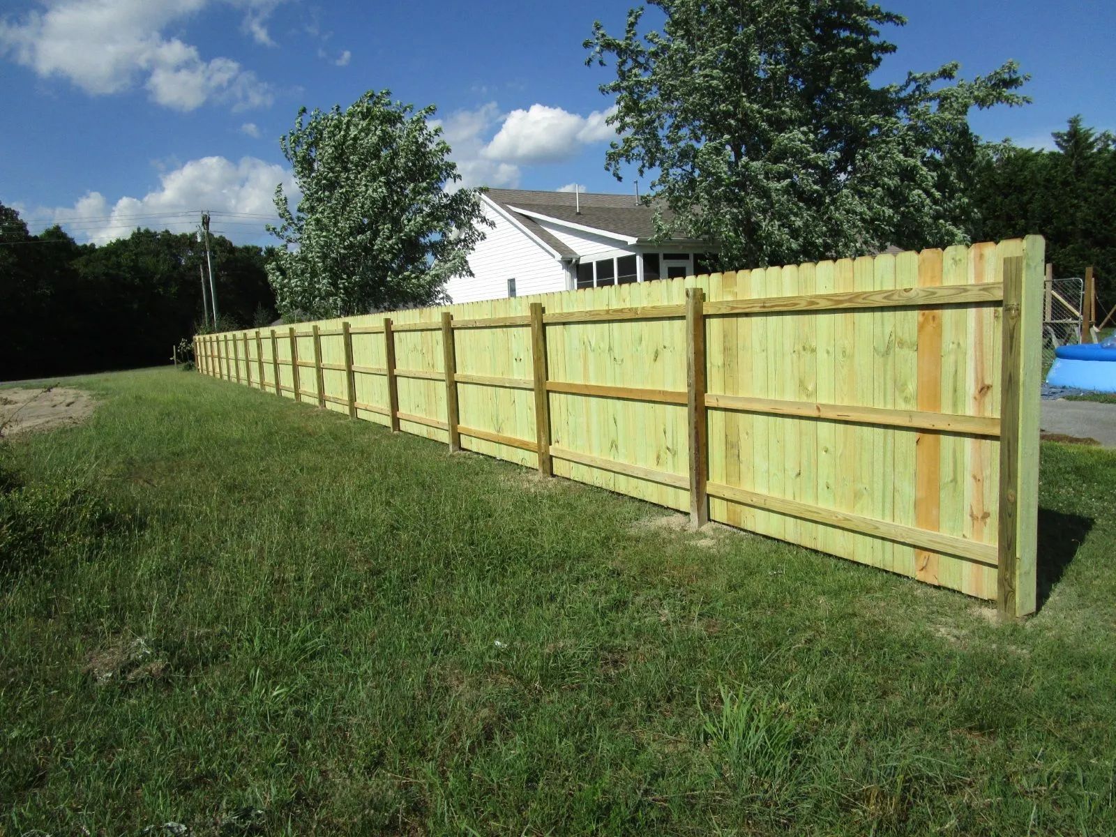 Long light wood fence along a grassy yard beside a house under a blue sky