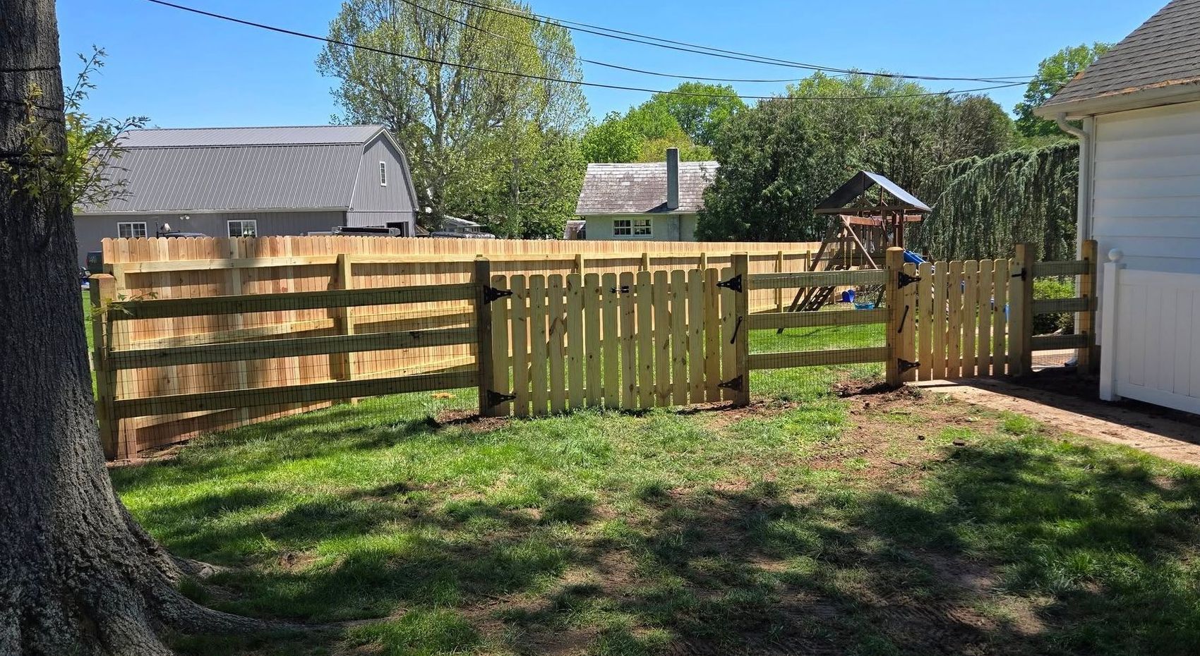 Wooden fence with gates in a grassy backyard. A swing set is visible beyond the fence, with houses in the background.