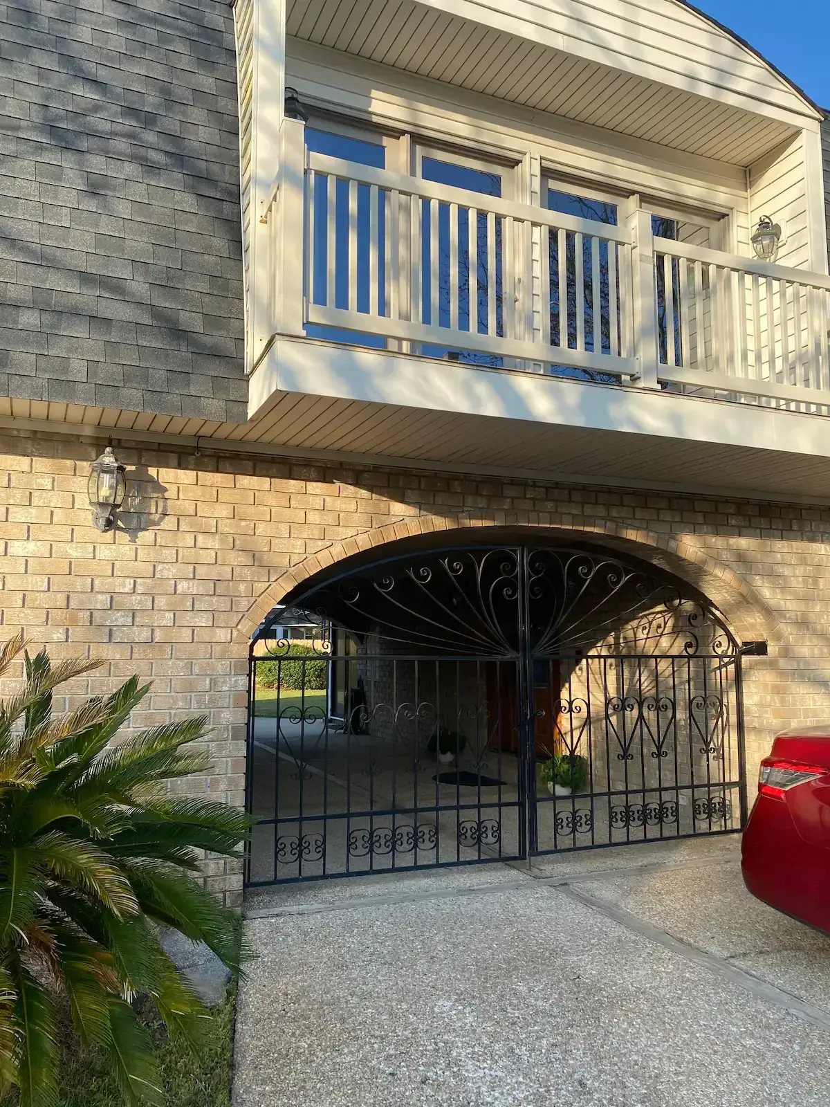 A red car is parked in front of a brick house with a balcony.