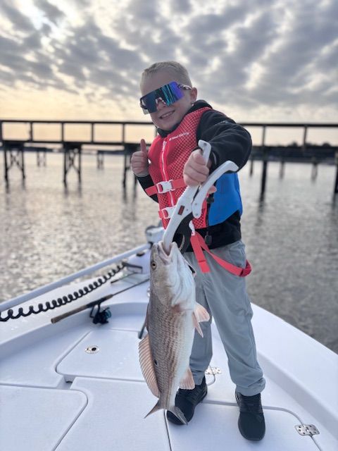A young boy on a boat holding a fish and giving a thumbs up