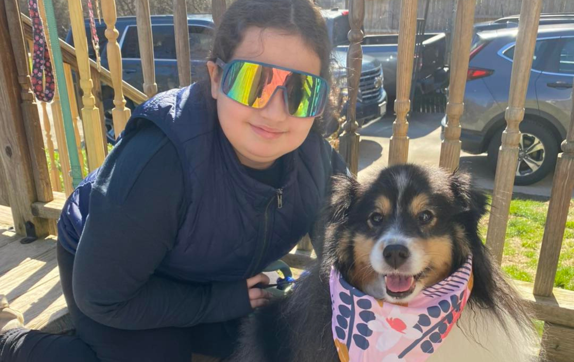 Person wearing sunglasses and vest with dog wearing a bandana, outside on a porch.