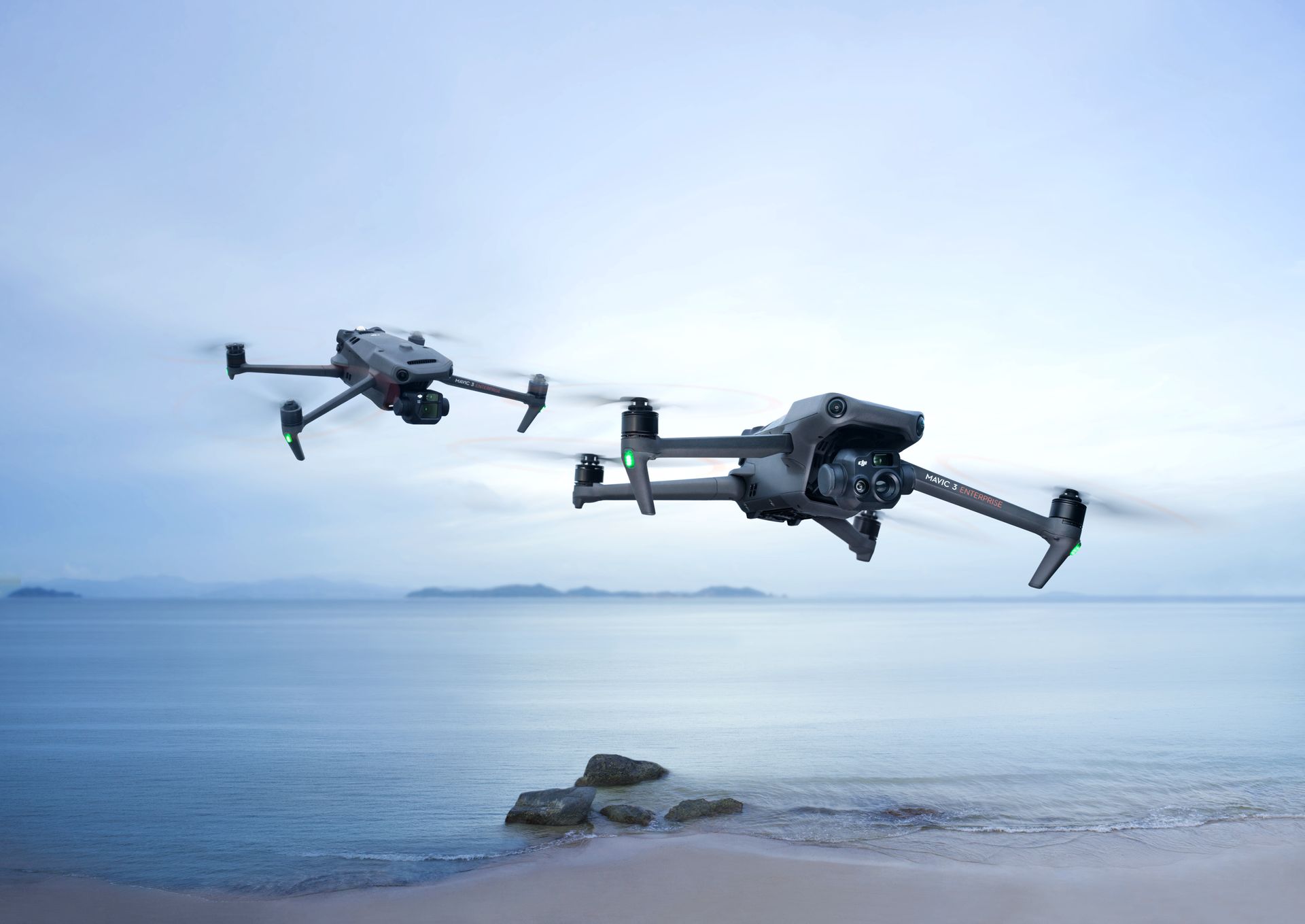 Two drones are flying over a beach near the ocean.